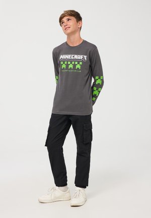 Maglietta grigia a maniche lunghe con logo di Minecraft in bianco e design pixelati verdi sulle maniche, abbinata a pantaloni cargo neri e sneakers bianche.