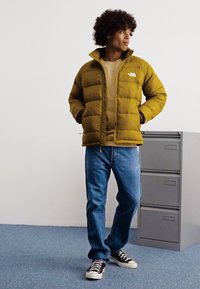 Sennepsgul puffjakke, beige skjorte, blå jeans, sort og hvidt sneakers. Jakken har lynlås og et logo på brystet.