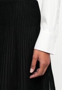 Fustă plisată neagră cu nervuri verticale, asociată cu o cămașă albă cu butoni. Mână odihnindu-se pe talia fustei.