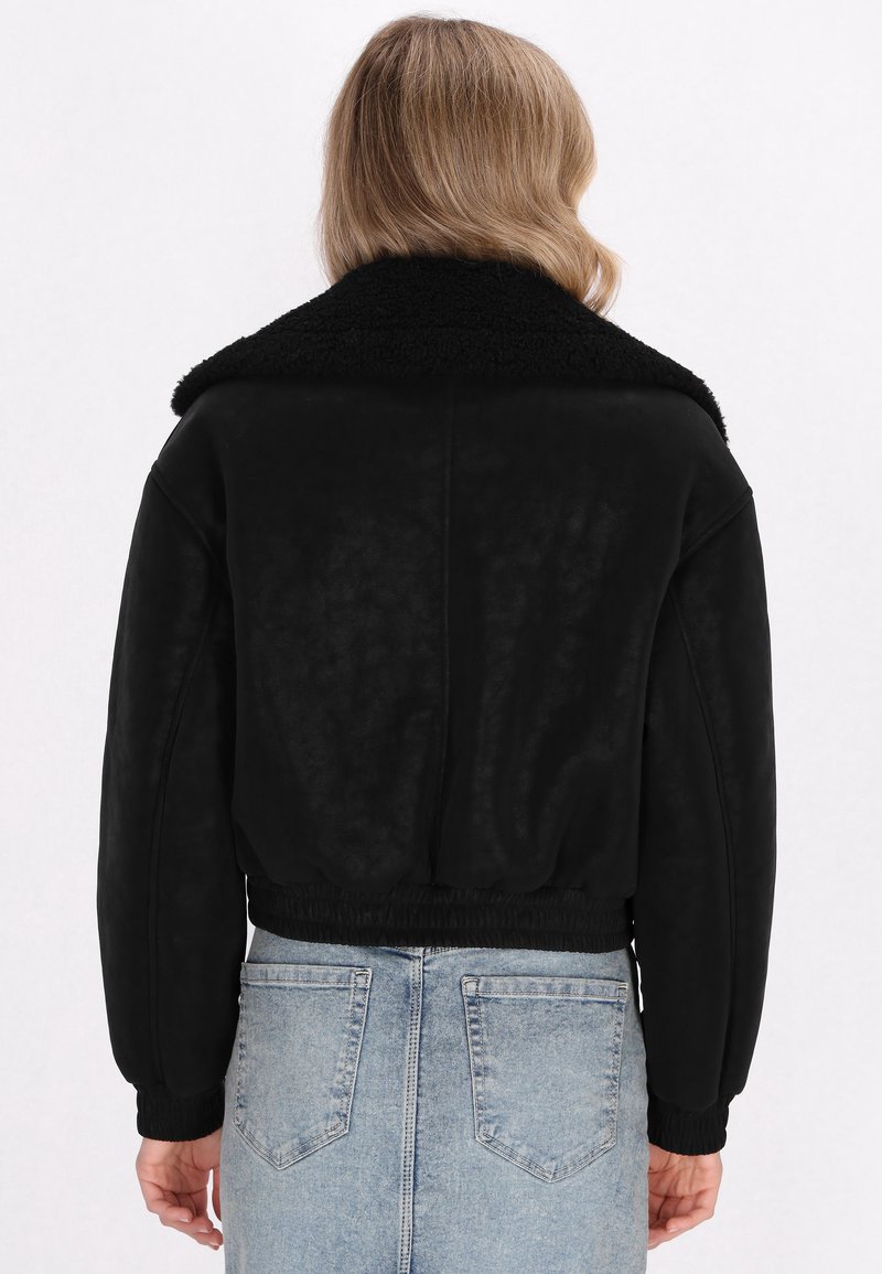 Personne avec des cheveux châtain clair de longueur épaules portant une veste en shearling noire et un jean taille haute bleu clair, de dos.