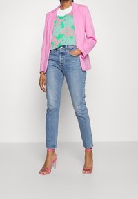 Blazer rosa ajustado sobre una blusa de patrón verde y rosa, combinado con pantalones vaqueros de color azul claro y sandalias de tacón alto rosa con tiras.