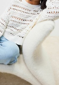 Personne portant un pull en crochet blanc et un jean bleu clair assise sur une chaise blanche et duveteuse incurvée sur un fond neutre.