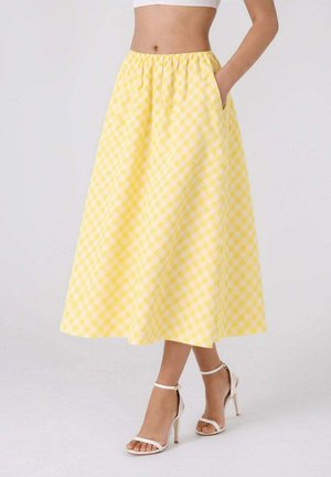 Femme portant une jupe midi vichy jaune avec des poches, associée à des sandales blanches à talons hauts avec bride à la cheville.