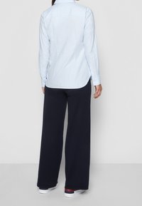 Camicia azzurra a maniche lunghe con orlo curvo e polsini bottonati, abbinata a pantaloni larghi blu scuro e sneakers bianche con accenti rossi.