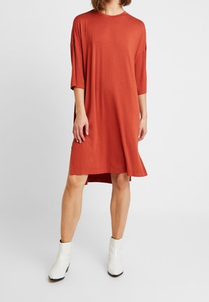 Kobieta w luźnej, rdzawej sukience do kolan z rękawami do łokcia i białych botkach, stojąca na białym tle.