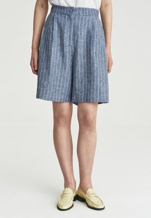 Person trägt hoch taillierte blaue Shorts mit weißen vertikalen Streifen und blassgelben Loafers und steht vor einem schlichten hellen Hintergrund.