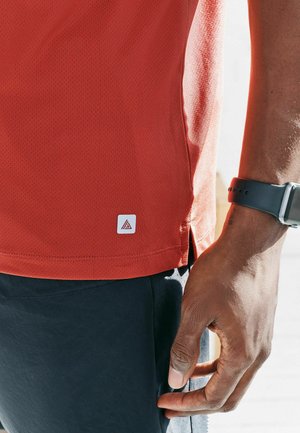 Close-up of a person’s hand wearing a black smartwatch next to a red athletic shirt and dark shorts.