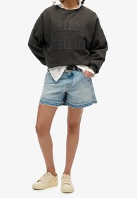 Szara bawełniana bluza z napisem "SUPERDRY ATHLETIC", zestawiona z niebieskimi szortami dżinsowymi z postrzępionymi krawędziami i jasnym praniem. Całość dopełniają beżowe sneakersy.