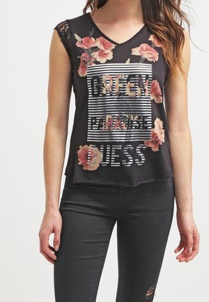 Kobieta nosząca czarny kwiatowy top bez rękawów z koronkowymi ramionami i paskowanym napisem "Green Paradise Guess,", w połączeniu z czarnymi podartymi jeansami.