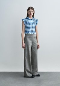 Kvinde står iført en blå og hvidternet ærmeløs bluse med flæser og højtaljede, vidde grå bukser og sorte sko.