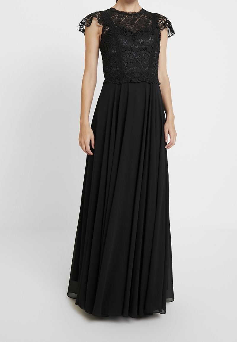 Femme portant une robe longue noire avec des manches courtes en dentelle et un corsage en dentelle florale, debout devant un fond blanc uni.