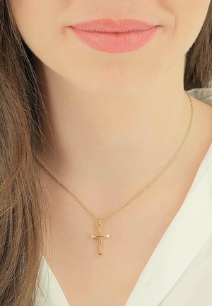 Collier en or avec un petit pendentif croix, présentant un design simple et une texture lisse, porté contre une chemise de couleur claire.