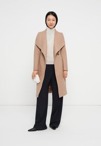Frau steht in einem beigen langen Mantel über einem weißen Rollkragenpullover, schwarzen weiten Hosen, schwarzen Schuhen und hält eine kleine weiße Clutch.