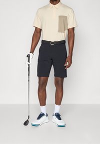 Tenue de golf pour homme comprenant un polo beige à manches courtes avec une poche beige et un short bleu marine. Chaussures blanches avec des accents bleus, tenant un club de golf.