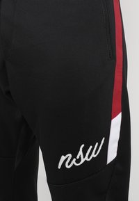 Zwarte joggers met rode zijkanten, voorzien van een witte geborduurde logo. Gemaakt van een gladde, elastische stof met ritszakken.