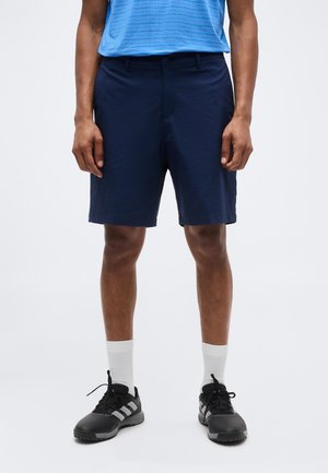 Homme portant un short bleu marine, une chemise à rayures bleues, des chaussettes blanches et des chaussures de sport noires, debout devant un fond uni.