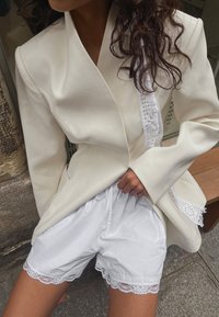 Blazer blanc ajusté avec un col texturé ; associé à un short blanc avec une bordure en dentelle. Tissus doux, coupe décontractée et design superposé.