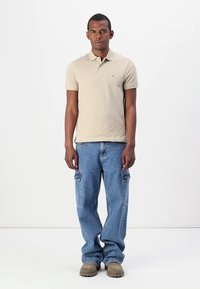 Béžové polo triko s texturovaným vzorem, spárované se světle modrými volnými džíny s cargo kapsami. Hnědé obuv dotváří celý outfit.