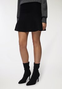 Donna che indossa una mini gonna nera a campana, un maglione grigio a maniche lunghe e stivaletti neri con tacco a punta, in piedi su uno sfondo bianco.