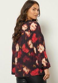 Donna con lunghi capelli castani che indossa una blusa nera a maniche lunghe e larghe con un motivo floreale rosso e bianco, girata a guardare oltre la spalla.