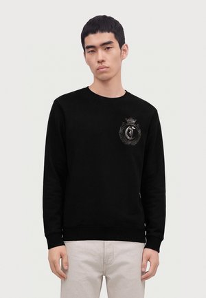 Young man wearing black sweatshirt with dark emblem on the left chest, standing against a plain light background.