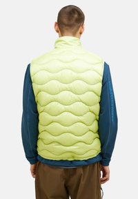 Gepolsterte, limegrüne Weste mit Wellenmuster, hohem Kragen und glatter Textur, über einem blauen Langarmshirt getragen.