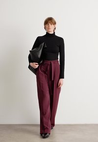 Svart turtleneck topp parret med høy midje, vide burgunder bukser. Modellen holder en svart strukturert håndveske, med enkle svarte sko.