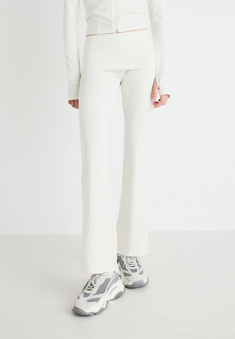 Leggings bianchi aderenti con fondo svasato, design senza cuciture, abbinati a sneaker grigie e bianche con suola spessa, su uno sfondo bianco uniforme.