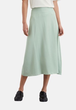 Vrouw draagt een lichtgroene midi-rok, wit topje en zwarte loafers, staat met licht gespreide benen tegen een effen achtergrond.
