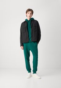 Svart zip-up jakke over en grønn hettegenser og matchende joggebukser. Hvite sneakers, frontlommer på jakken, minimalistisk design.