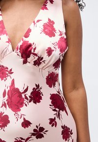 Κοντινή λήψη ενός αμάνικου ανοιχτό ροζ φορέματος με βαθύ V λαιμόκοψη και σκούρο ροζ λουλουδάτο σχέδιο σε άτομο με μεσαίο τόνο επιδερμίδας.