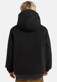 Crna oversized jakna s velikom kapuljačom, elastičnim manžetama i rebrastim donjim dijelom. Materijal se čini glatkima i čvrstim. Nema vidljivih uzoraka ili akcenata.