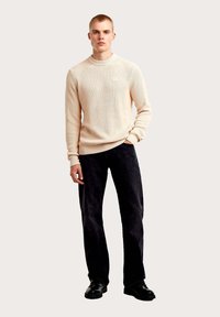 Beige geribbelde trui met een ronde hals, gecombineerd met zwarte broek met wijde pijpen en zwarte schoenen. De trui heeft een klein logo op de borst.