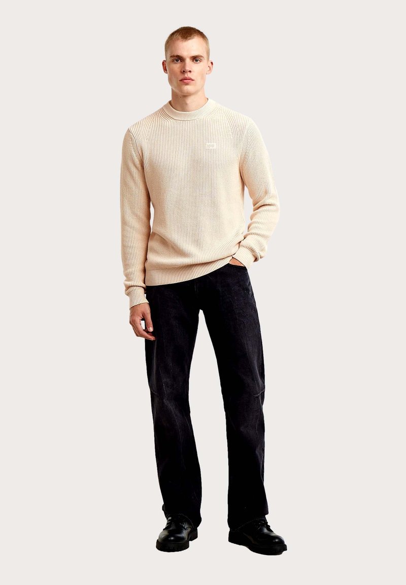 Jersey beige acanalado con cuello redondo, combinado con pantalones anchos negros y zapatos negros. El jersey presenta un pequeño logo en el pecho.