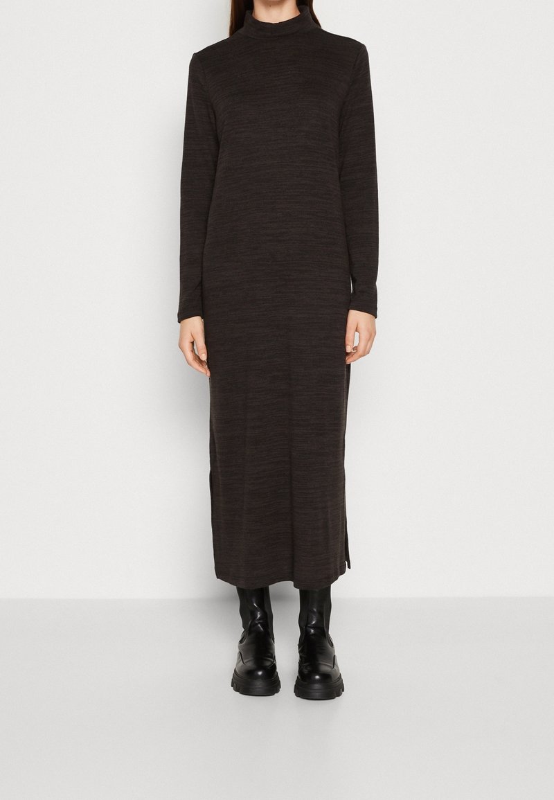 Femme portant une robe longue marron foncé à manches longues avec col haut et des bottes noires épaisses, debout sur un fond clair.