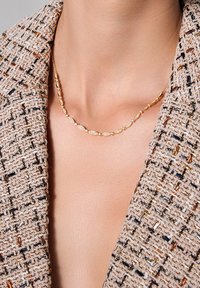 Złoty naszyjnik z owalnymi ogniwami, przeplatany małymi okrągłymi kamieniami. Noszony z fakturowaną beżową kurtką z ciemnymi akcentami.