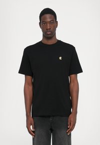 Tricou negru din bumbac, cu guler rotund, având un mic logo auriu pe piept și mâneci scurte, asociat cu blugi gri din denim.