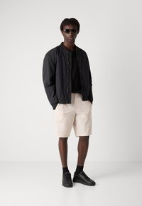 Svart bomberjacka över en svart t-shirt, beige shorts och svarta sneakers. Modell som bär mörka solglasögon, står mot en ljus bakgrund.