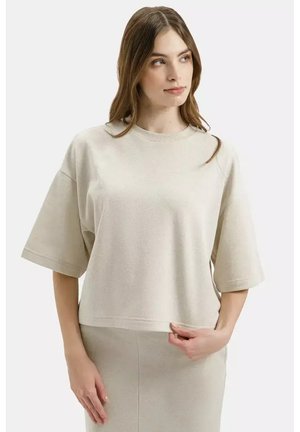 Kobieta ubrana w beżową, oversize'ową bluzkę z krótkim rękawem i dopasowaną spódnicę, patrząca w bok, trzymająca jedną ręką rąbek bluzki.