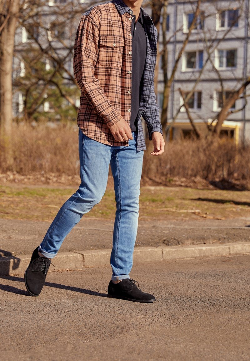 Personne marchant sur le trottoir portant une chemise à carreaux moitié marron, moitié grise, un t-shirt noir, un jean bleu clair et des chaussures noires.