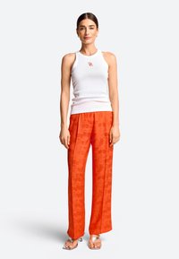 Weißes kurzärmeliges, geripptes Oberteil mit kleinem Logo, kombiniert mit orangen gemusterten, weit geschnittenen Hosen und silbernen Sandalen mit offenen Zehen.