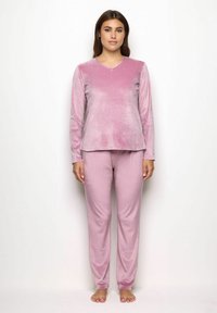 Donna in piedi, a piedi nudi, con un morbido top a maniche lunghe di velluto rosa e pantaloni abbinati, su uno sfondo bianco semplice.