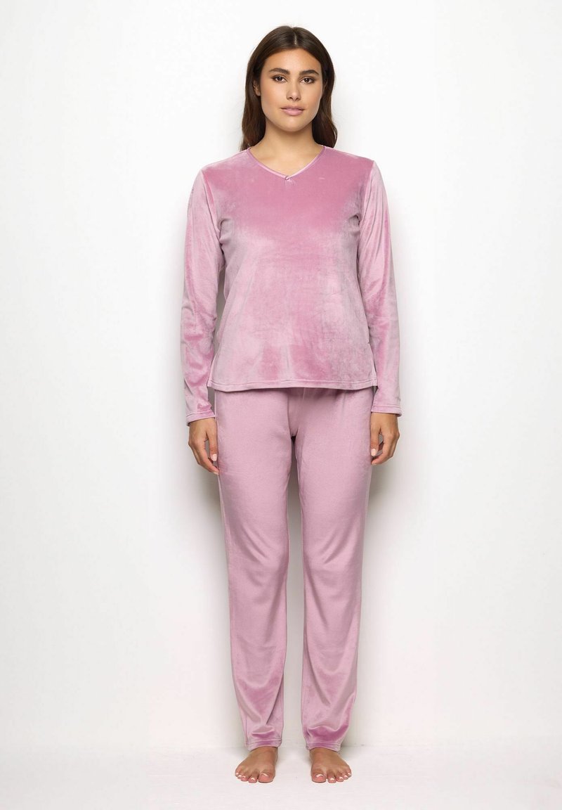 Donna in piedi, a piedi nudi, con un morbido top a maniche lunghe di velluto rosa e pantaloni abbinati, su uno sfondo bianco semplice.