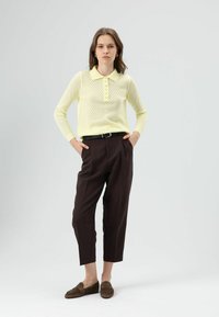 Gele gebreide polo met structuur en een kraag, voorzien van een knoopsluiting, gecombineerd met donkerbruine, hooggewaste broek en bruine suède loafers.