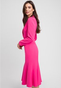 Dopasowana różowa sukienka z rozkloszowanym dołem, długimi rękawami i dopasowanym krojem. Gładka tekstura tkaniny podkreśla intensywny kolor.