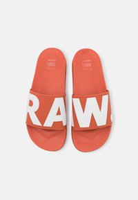 Red slide sandals featuring a wide white strap with the text "RAW." Smooth rubber texture with a flat sole and minimal branding on the footbed.