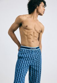 Blauwe pyjamabroek met een herhalend geometrisch ontwerp, voorzien van een witte Lacoste-logo tailleband, losse pasvorm en ademende stof.
