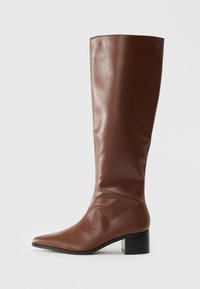 Botte en cuir marron jusqu'au genou avec un bout pointu et un talon bloc sur un fond blanc.