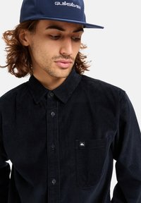 Navy corduroy button-up shirt with a small chest pocket and logo patch. Features a collar and vertical ribbed texture.