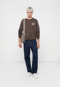 Brown sweatshirt with white "GAP" logo, blue straight-leg jeans, and white sneakers. Gray shoulder bag rests on left shoulder. Plain background.